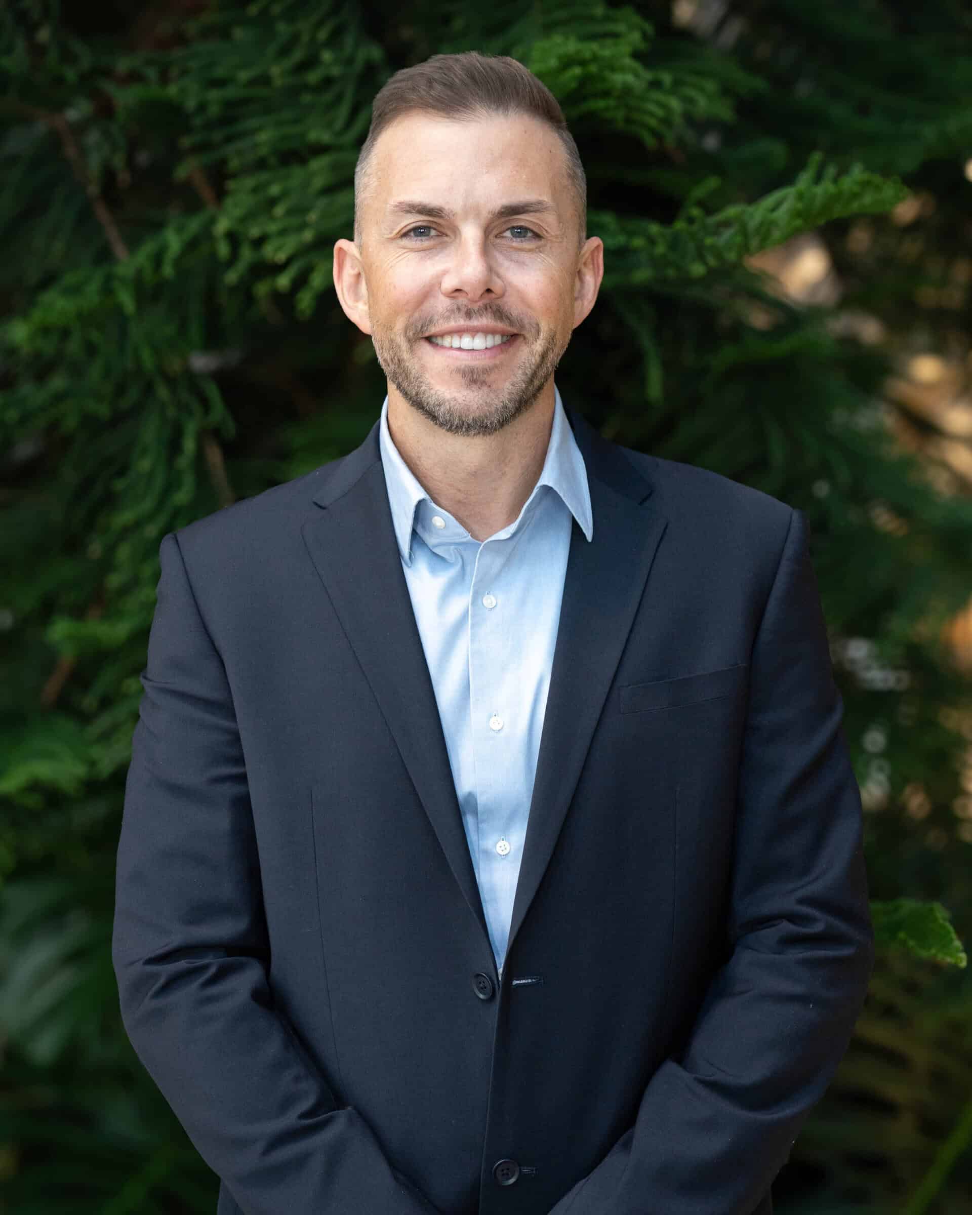 Professional headshot of a man wearing a light blue dress shirt and navy, standing in front of a tree.