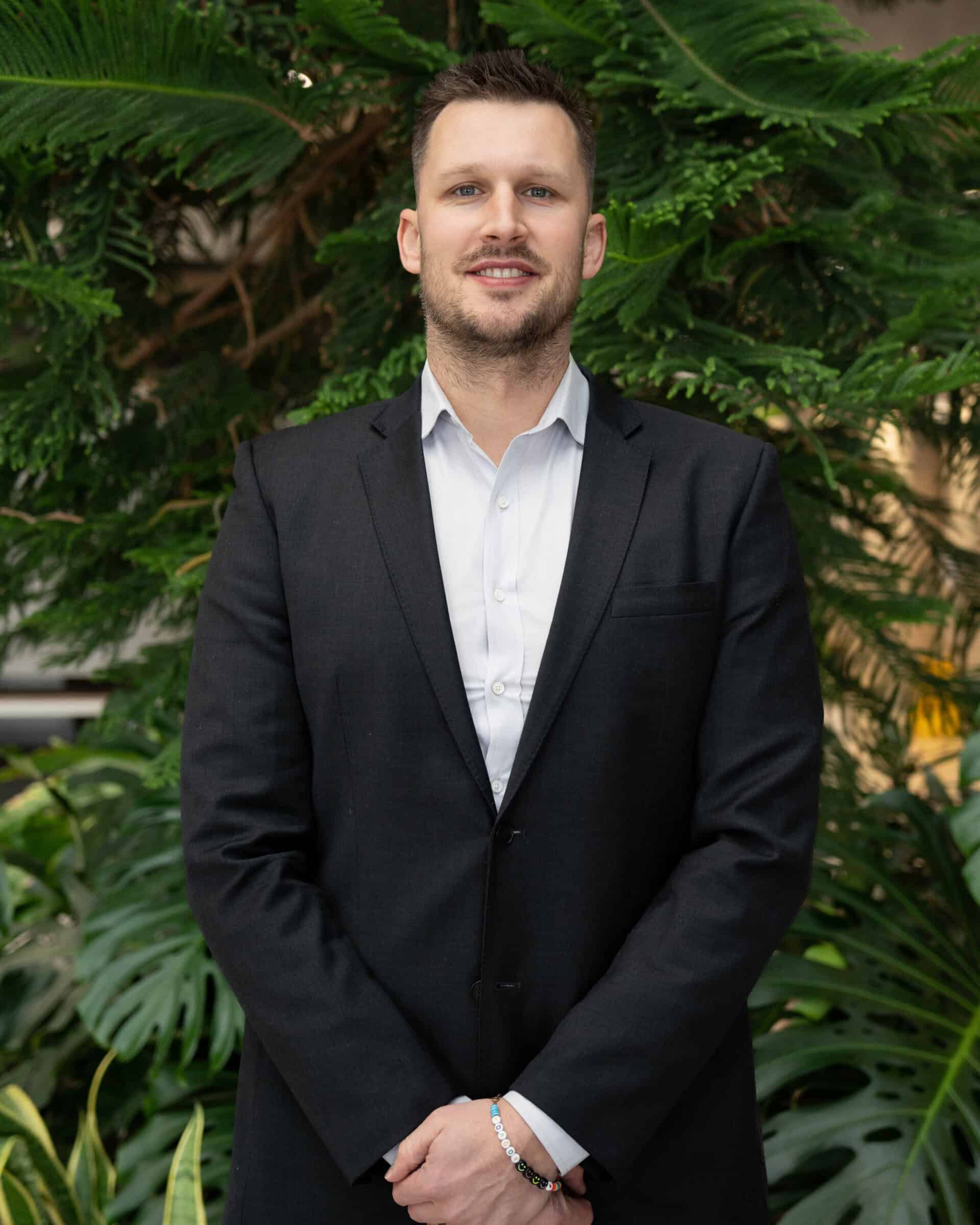 Professional headshot of a man wearing a white dress shirt and black blazer, standing in front of a tree.