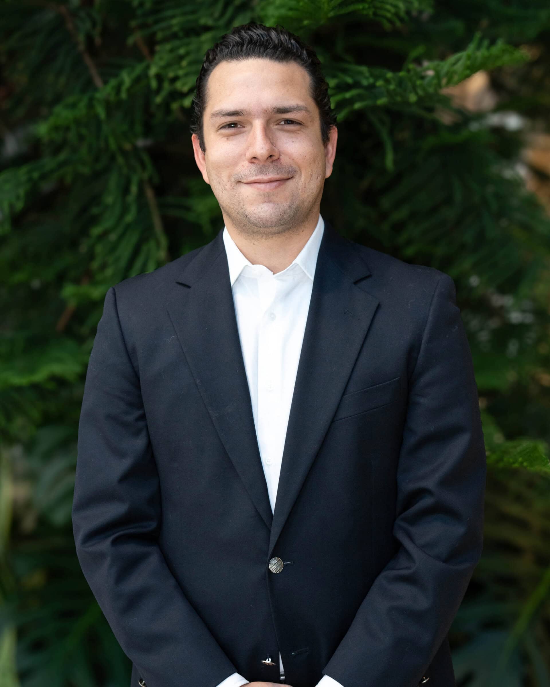 Professional headshot of a man wearing a white dress shirt and navy blazer, standing in front of a tree.
