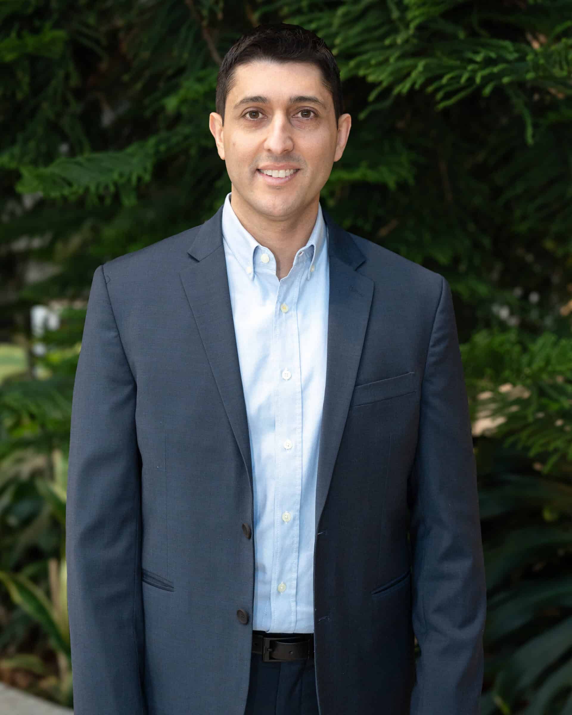 Professional headshot of a man wearing a light blue dress shirt and navy blazer, standing in front of a tree.