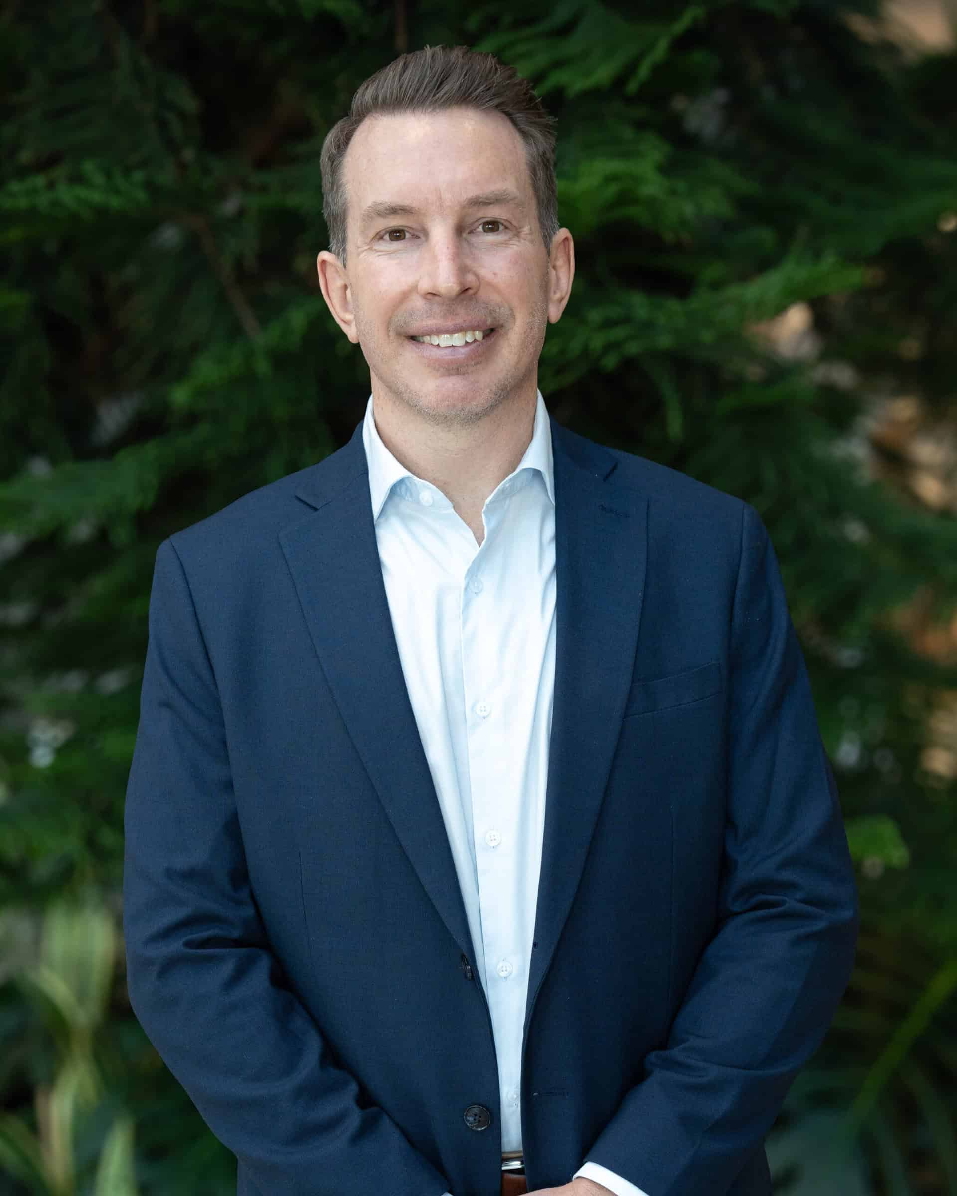 Professional headshot of a man wearing a white dress shirt and navy blazer, standing in front of a tree.