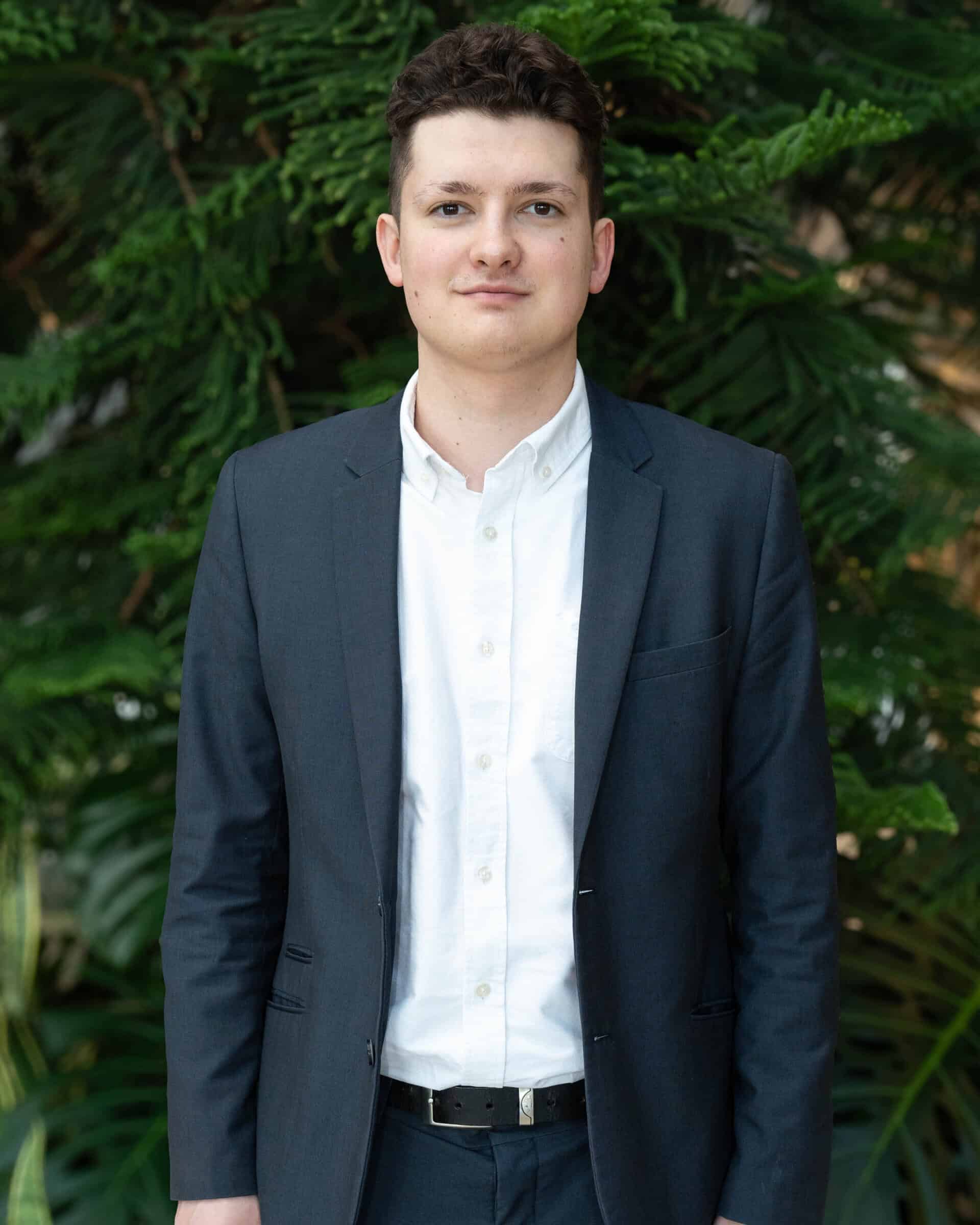 Professional headshot of a man wearing a white dress shirt and navy blazer, standing in front of a tree.