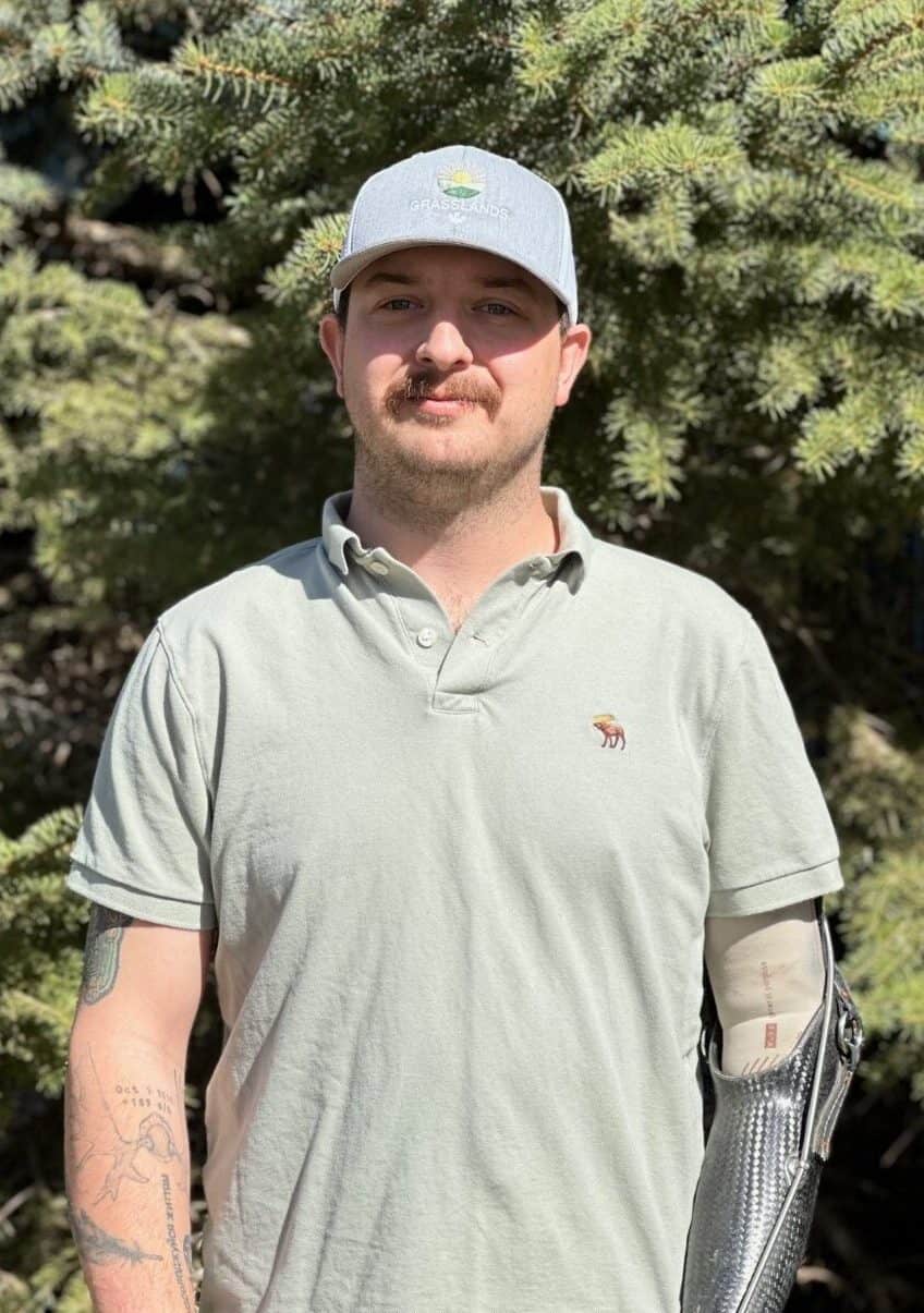 Lawn care professional outdoors wearing Grasslands hat and polo shirt with tree background.