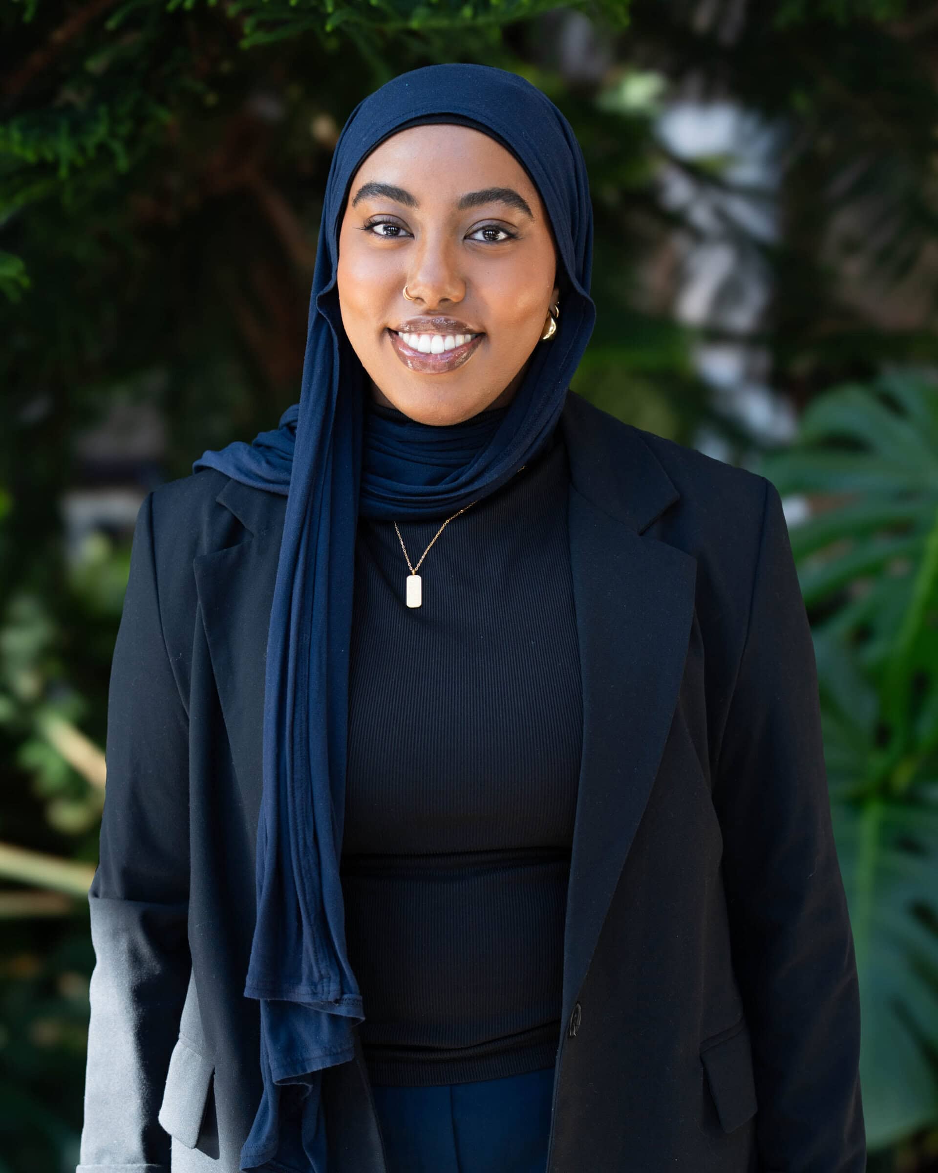 Professional headshot of a woman in a black shirt, black blazer, and navy blue hijab, standing in from t of a tree.