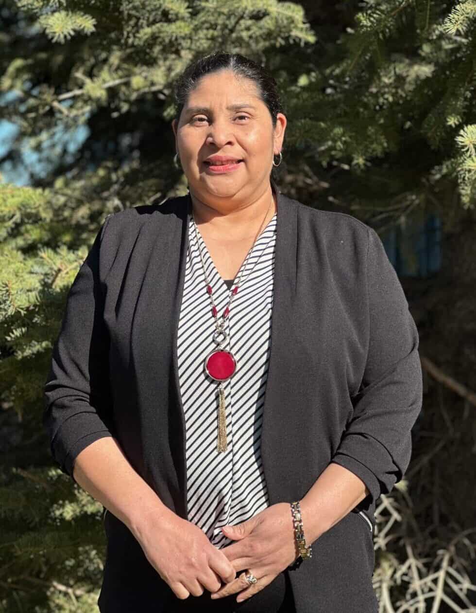 Professional headshot of a woman wearing a gray shit and black cardigan, standing in front of a tree.