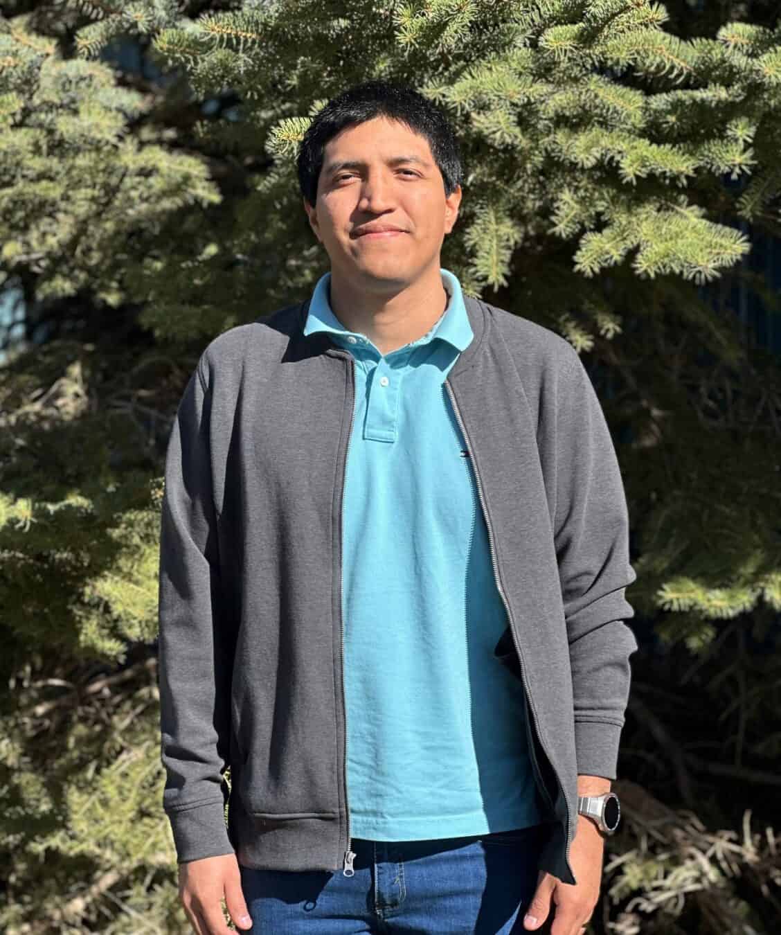 Professional headshot of a man in a blue shirt and gray sweater standing in front of a tree.
