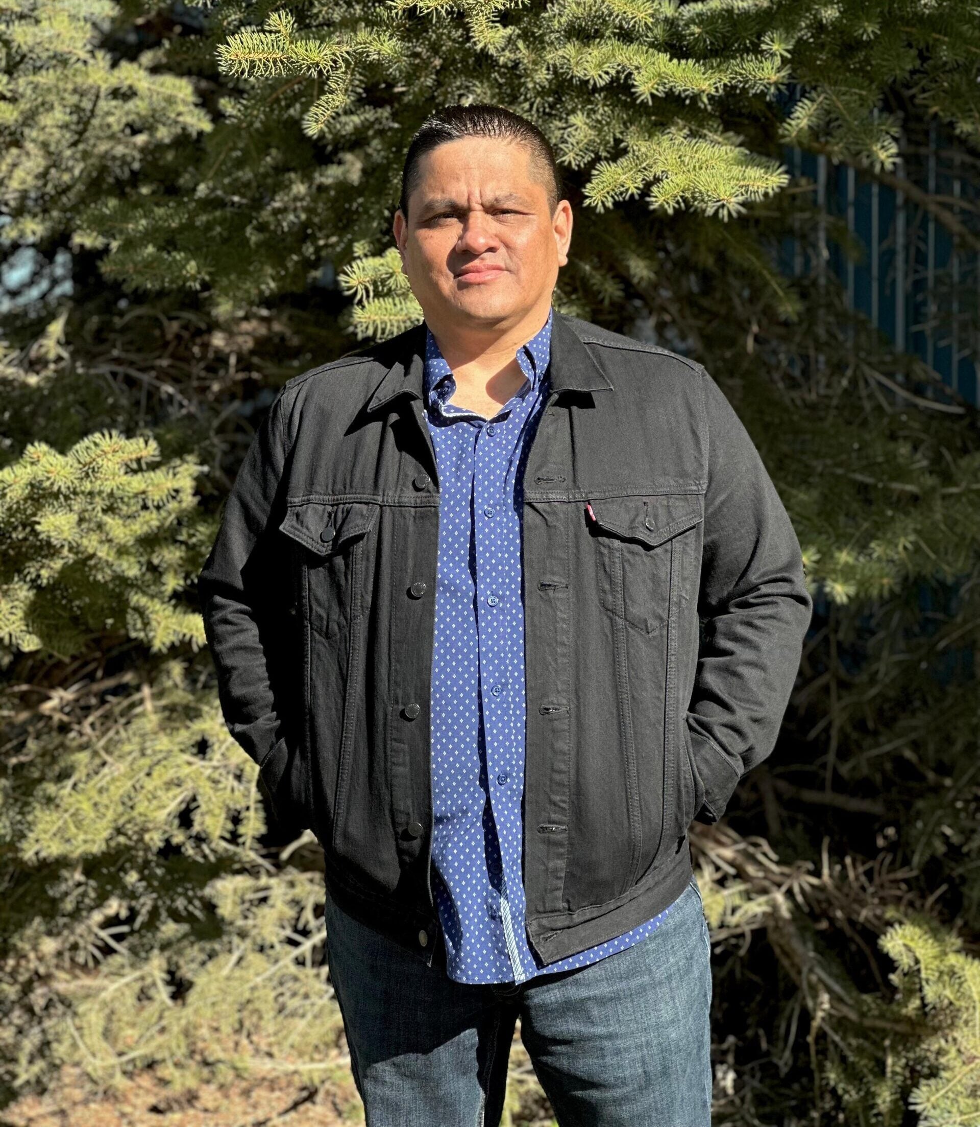 Professional headshot of a man in a blue shirt and black jean jacket standing in front of a tree.