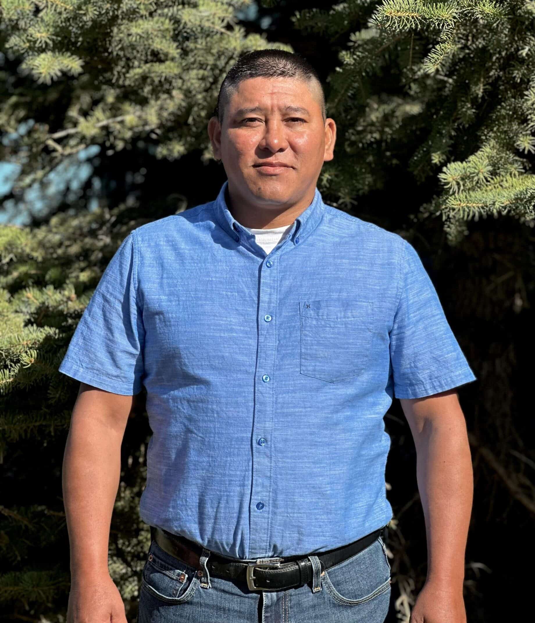 Professional headshot of a man in a blue button-up shirt, standing in front of a tree.