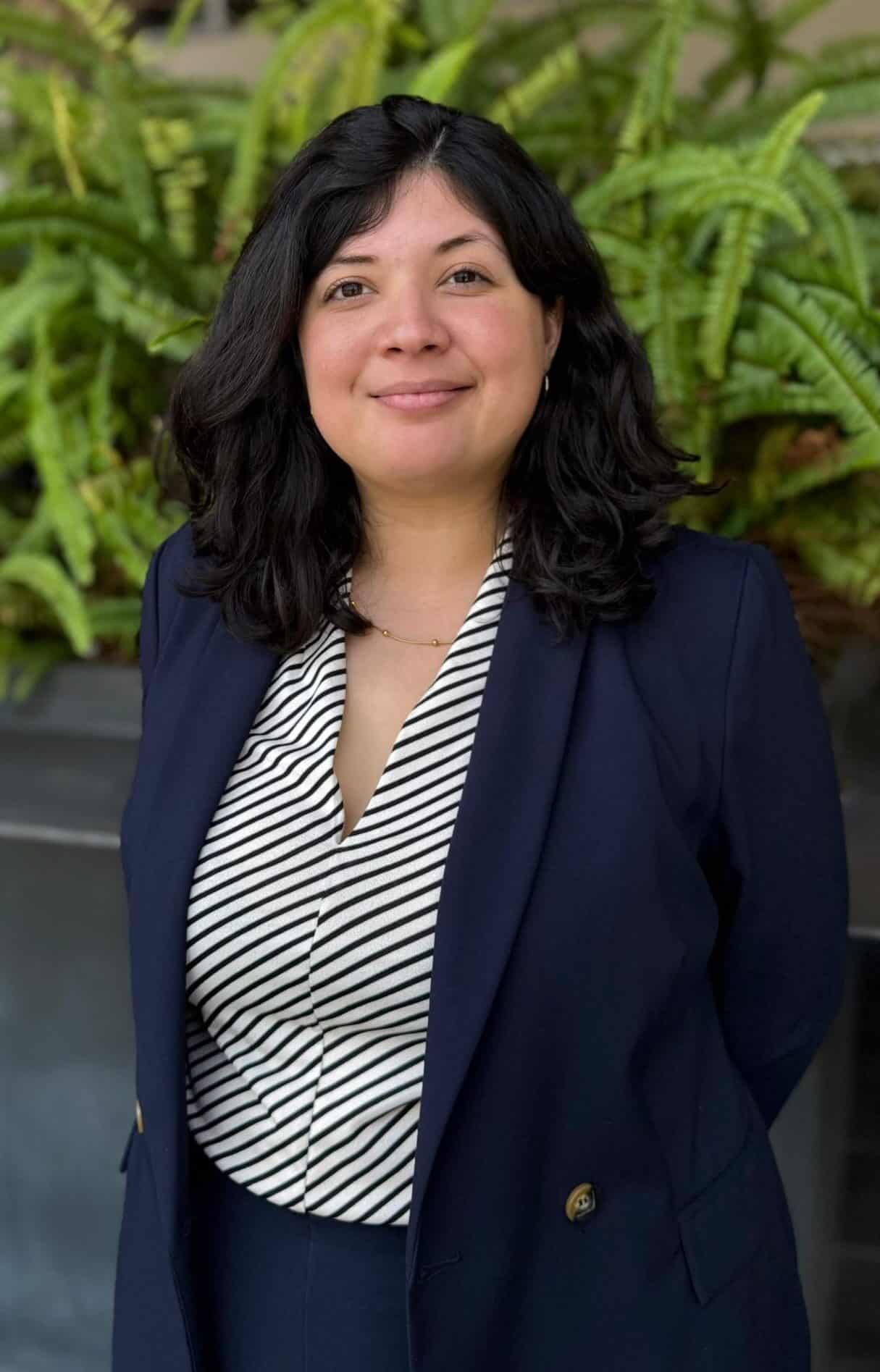 Healthy woman in business attire smiling outdoors with green plants in background.