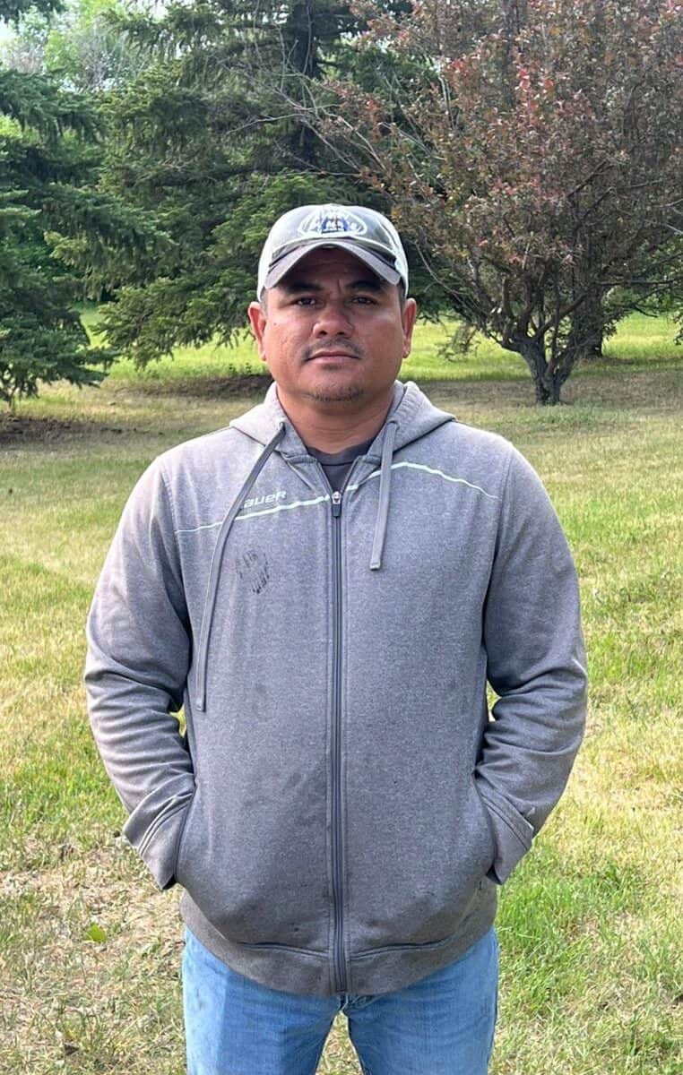 Man outdoors in a gray hoodie and cap, standing in a lush green park with trees.