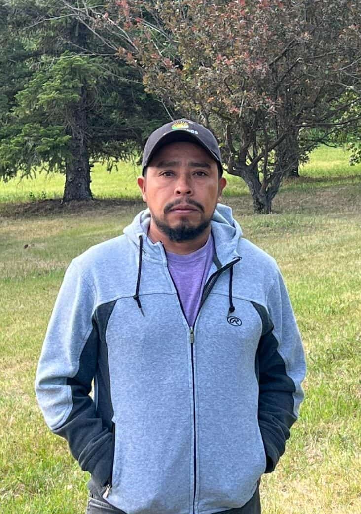 Man standing outdoors in a park with trees and grass, wearing casual hoodie and cap.