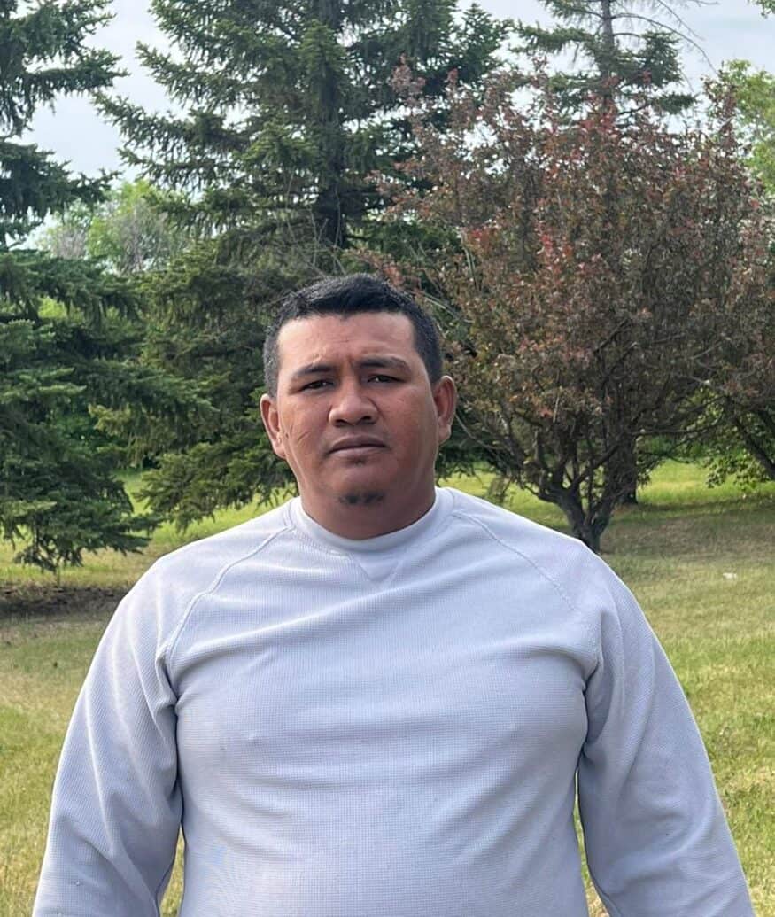 Man outdoors in a grassy park with trees, wearing a light gray sweatshirt.