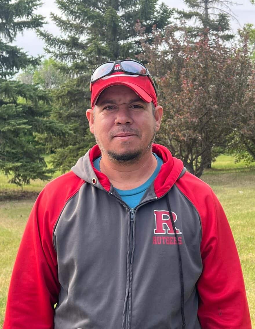 Man in Rutgers jacket standing outdoors among trees, casual sportswear, sunglasses on cap, natural greenery background.