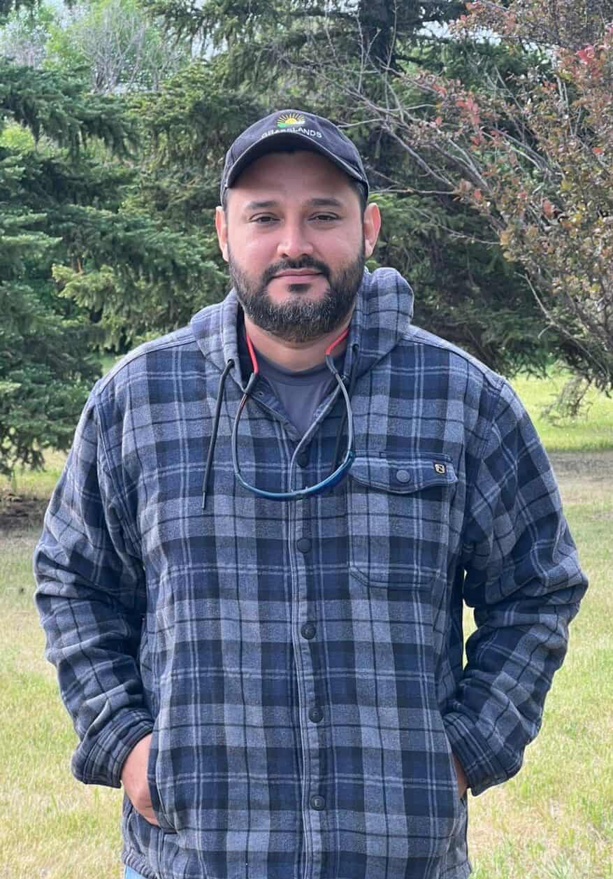 Man in outdoors wearing plaid jacket with trees in background.