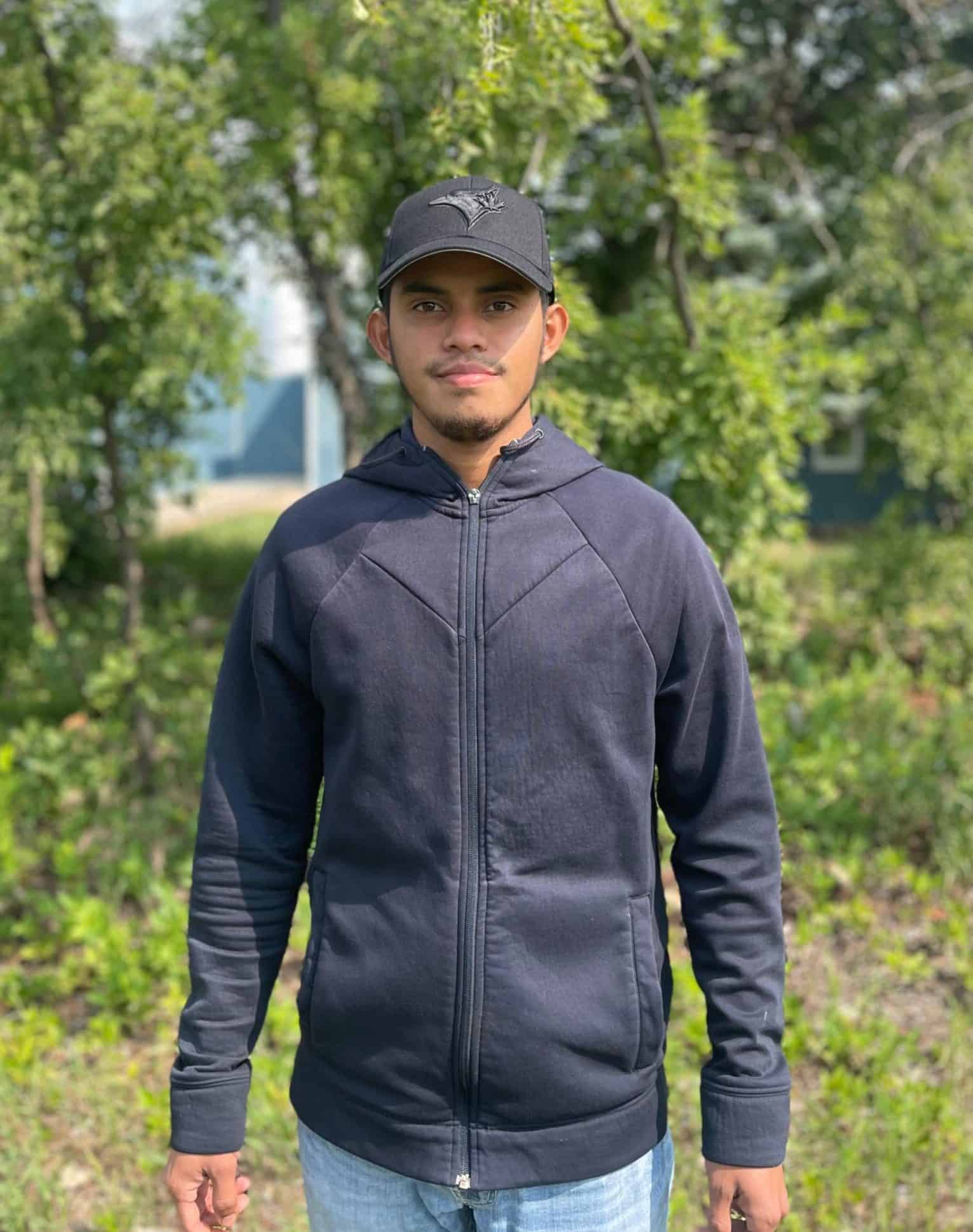 A young man wearing a black hoodie and cap standing outdoors in a green park.